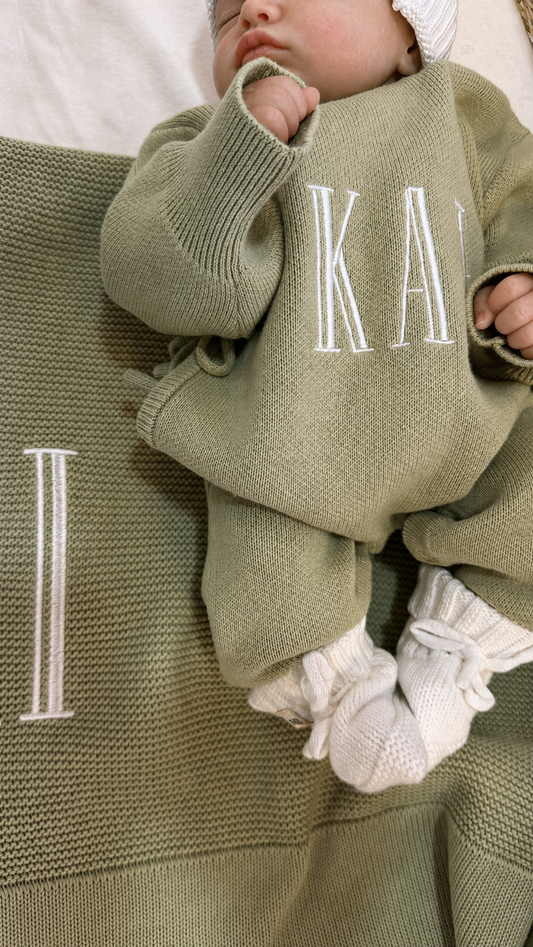 Close-up of name embroidery on a Soft Sage newborn knit romper with a matching blanket and Warm White booties.