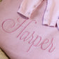 Close-up of a petal pink ruffle knit blanket embroidered with the name Harper and matching newborn accessories.