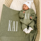 Newborn wearing a personalized Soft Sage knit romper with a Warm White hat and booties beside a matching name blanket.