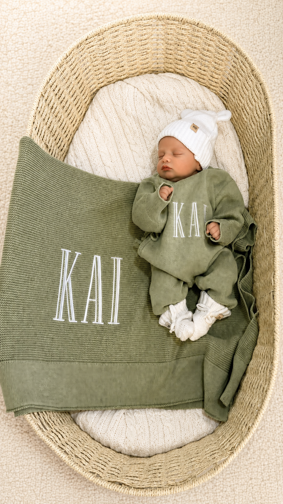 Newborn wearing a personalized Soft Sage knit romper with a Warm White hat and booties beside a matching name blanket.