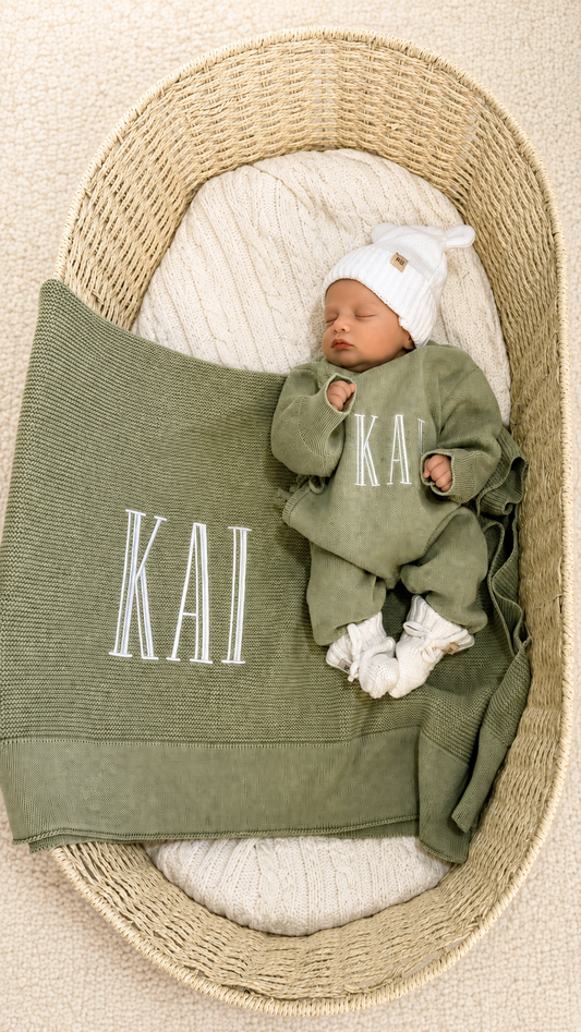 Newborn wearing a personalized Soft Sage knit romper with a Warm White hat and booties beside a matching name blanket.