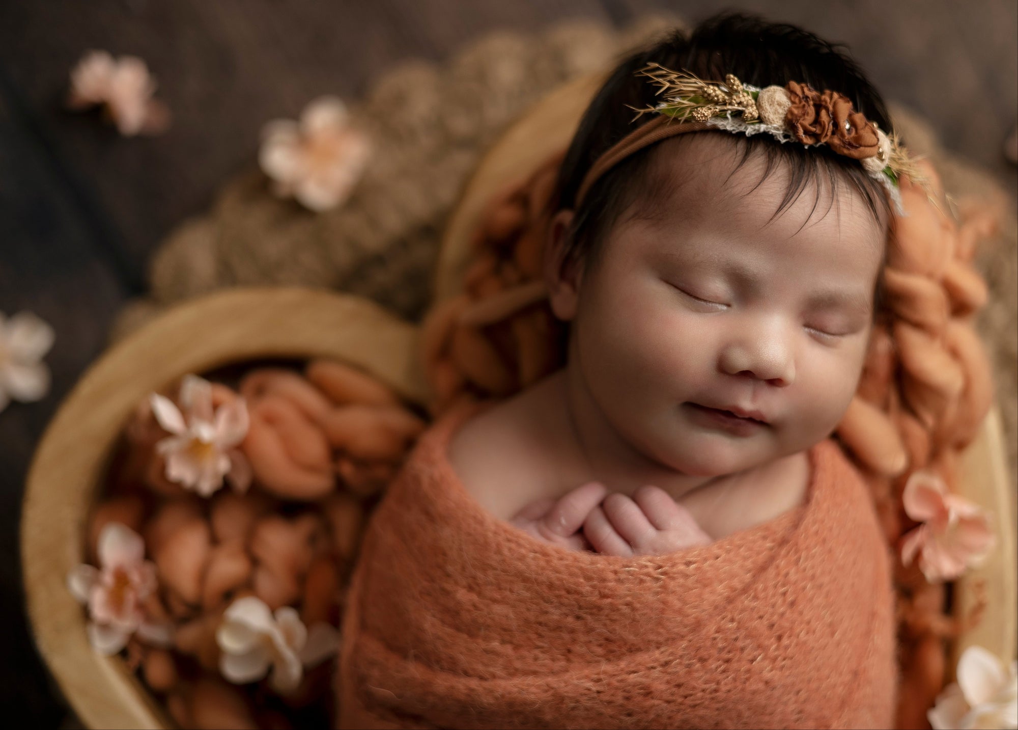 Newborn baby wrapped in a brown blanket with a floral headband, surrounded by flowers on a dark background.