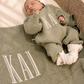 Newborn in a personalized Soft Sage knit romper with a matching name blanket, Warm White hat, and knit booties in a woven bassinet.