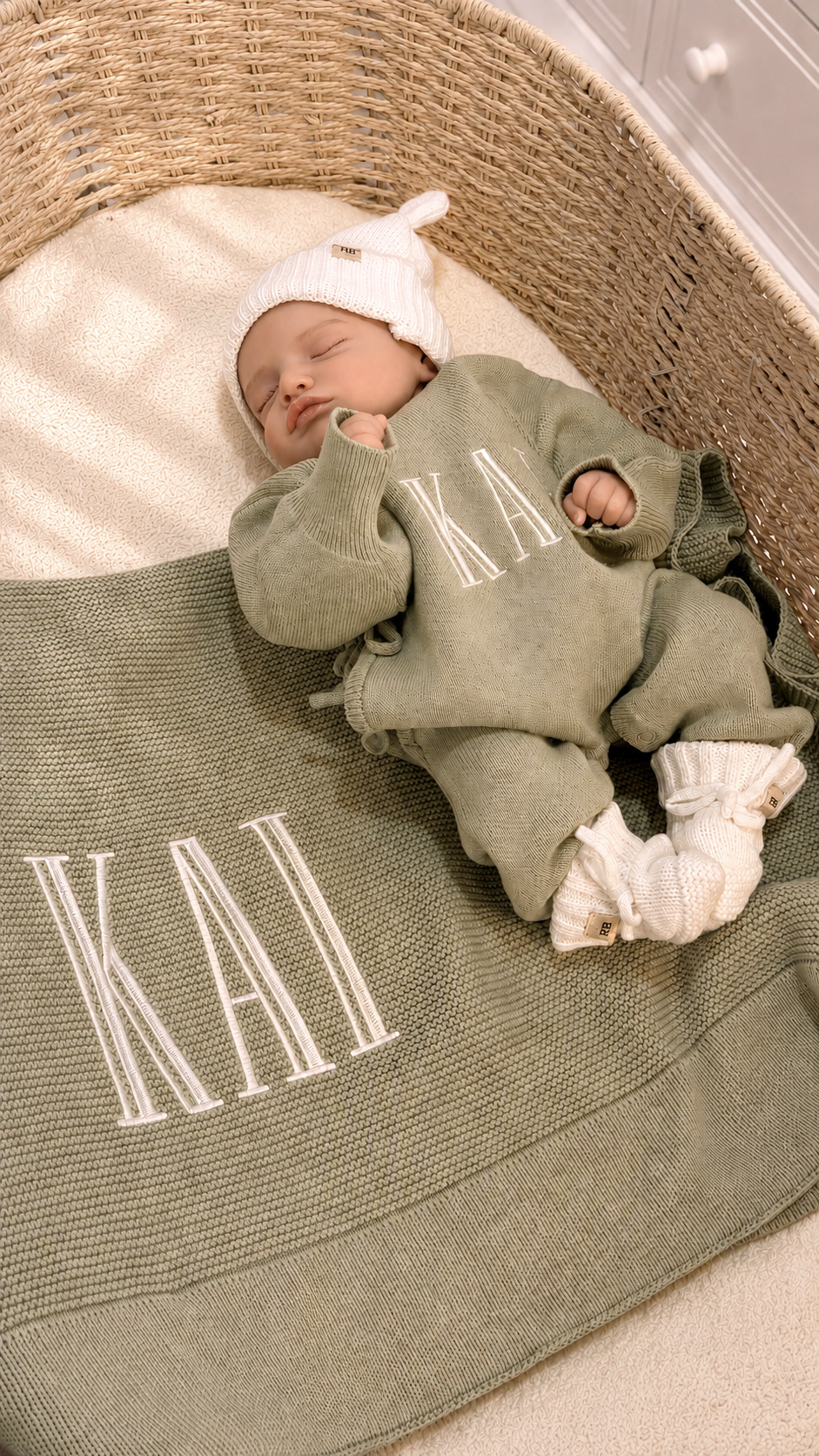 Newborn in a personalized Soft Sage knit romper with a matching name blanket, Warm White hat, and knit booties in a woven bassinet.