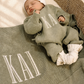 Newborn in a personalized Soft Sage knit romper with a matching name blanket, Warm White hat, and knit booties in a woven bassinet.