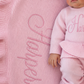 Newborn wearing a personalized petal pink Ballerina long romper beside a matching blanket embroidered with the name Harper.