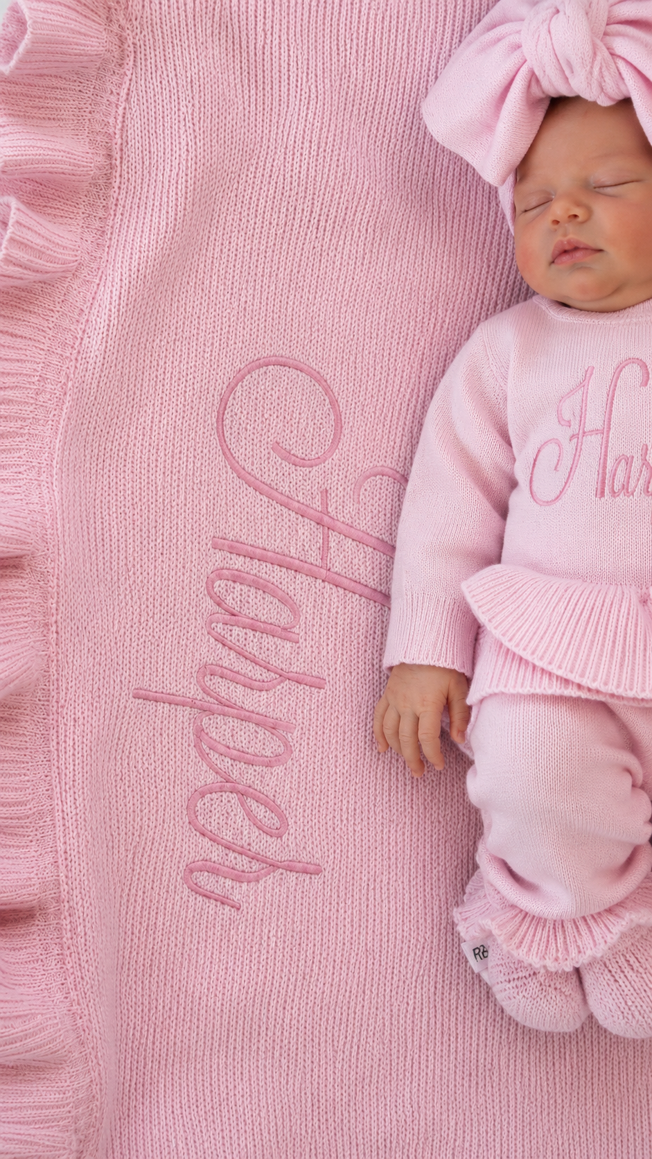 Newborn wearing a personalized petal pink Ballerina long romper beside a matching blanket embroidered with the name Harper.