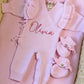 Personalized petal pink long newborn romper arranged with its matching bow and ruffle booties in a woven bassinet.