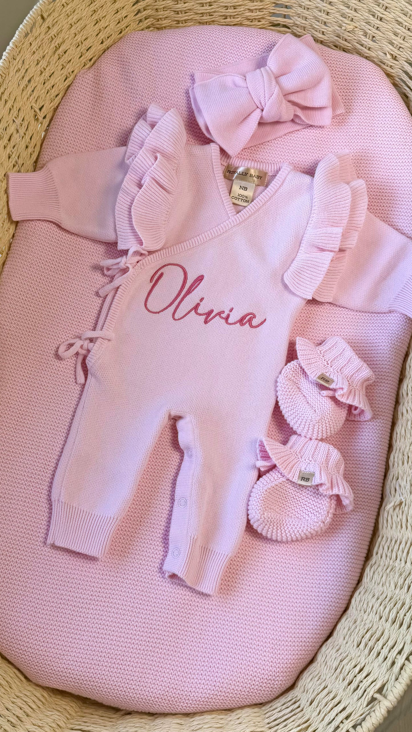 Personalized petal pink long newborn romper arranged with its matching bow and ruffle booties in a woven bassinet.