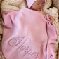 Newborn in a petal pink Ballerina long romper and bow headwrap, tucked beneath a personalized ruffle knit blanket in a bassinet.