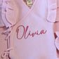 Close-up of personalized name embroidery and butterfly ruffle wrap detailing on a petal pink newborn romper.