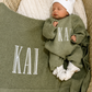 Newborn wearing a personalized Soft Sage knit romper with a Warm White hat and booties beside a matching name blanket.