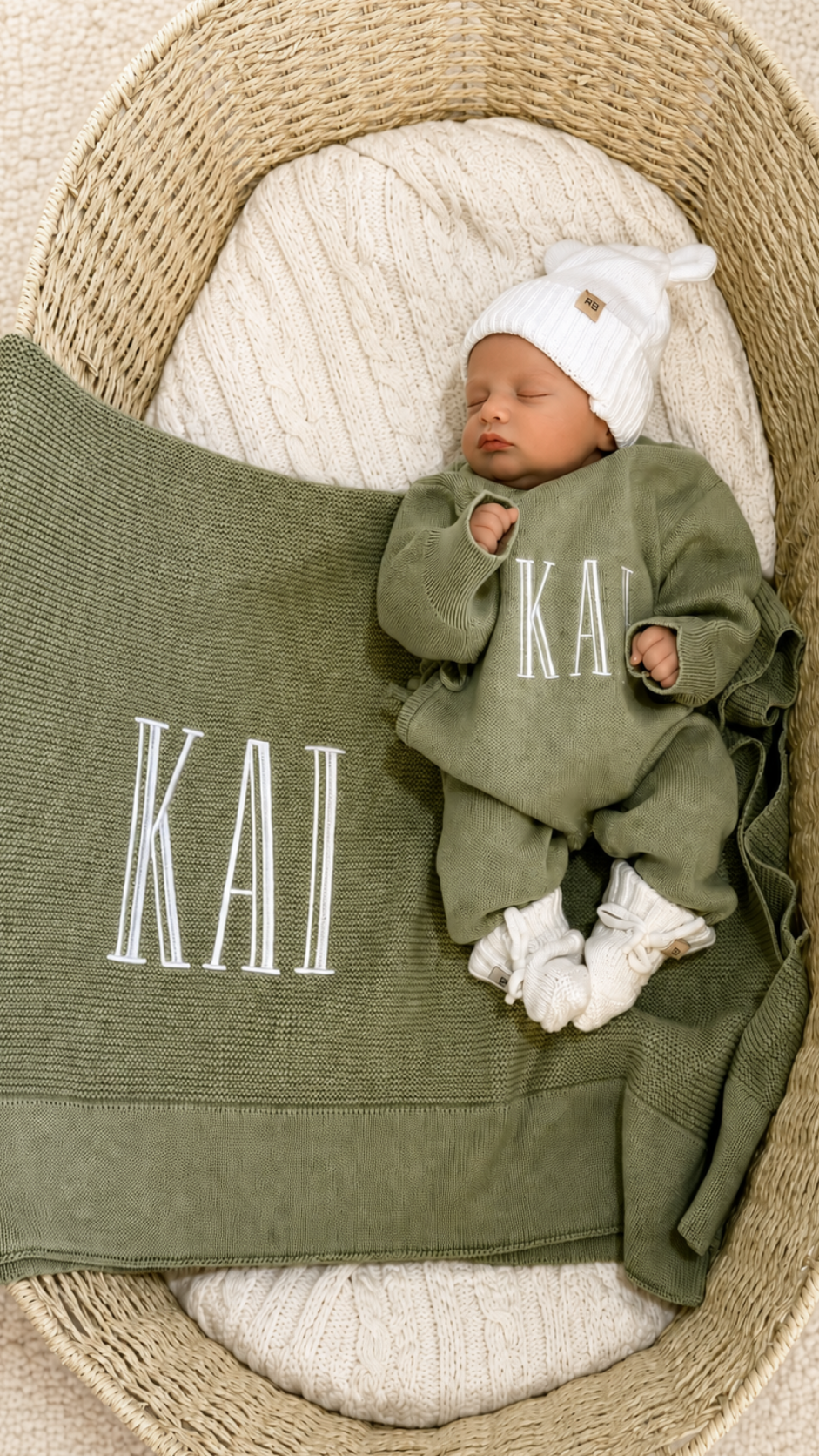 Newborn wearing a personalized Soft Sage knit romper with a Warm White hat and booties beside a matching name blanket.