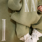 Close-up of name embroidery on a Soft Sage newborn knit romper with a matching blanket and Warm White booties.