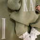 Close-up of name embroidery on a Soft Sage newborn knit romper with a matching blanket and Warm White booties.