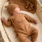 Newborn wearing a personalized Teddy Brown kimono romper beside the matching name blanket, bear-ear hat, and booties