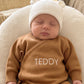 Newborn wearing a personalized Teddy Brown knit romper with a Warm White bear-ear hat and matching knit booties.