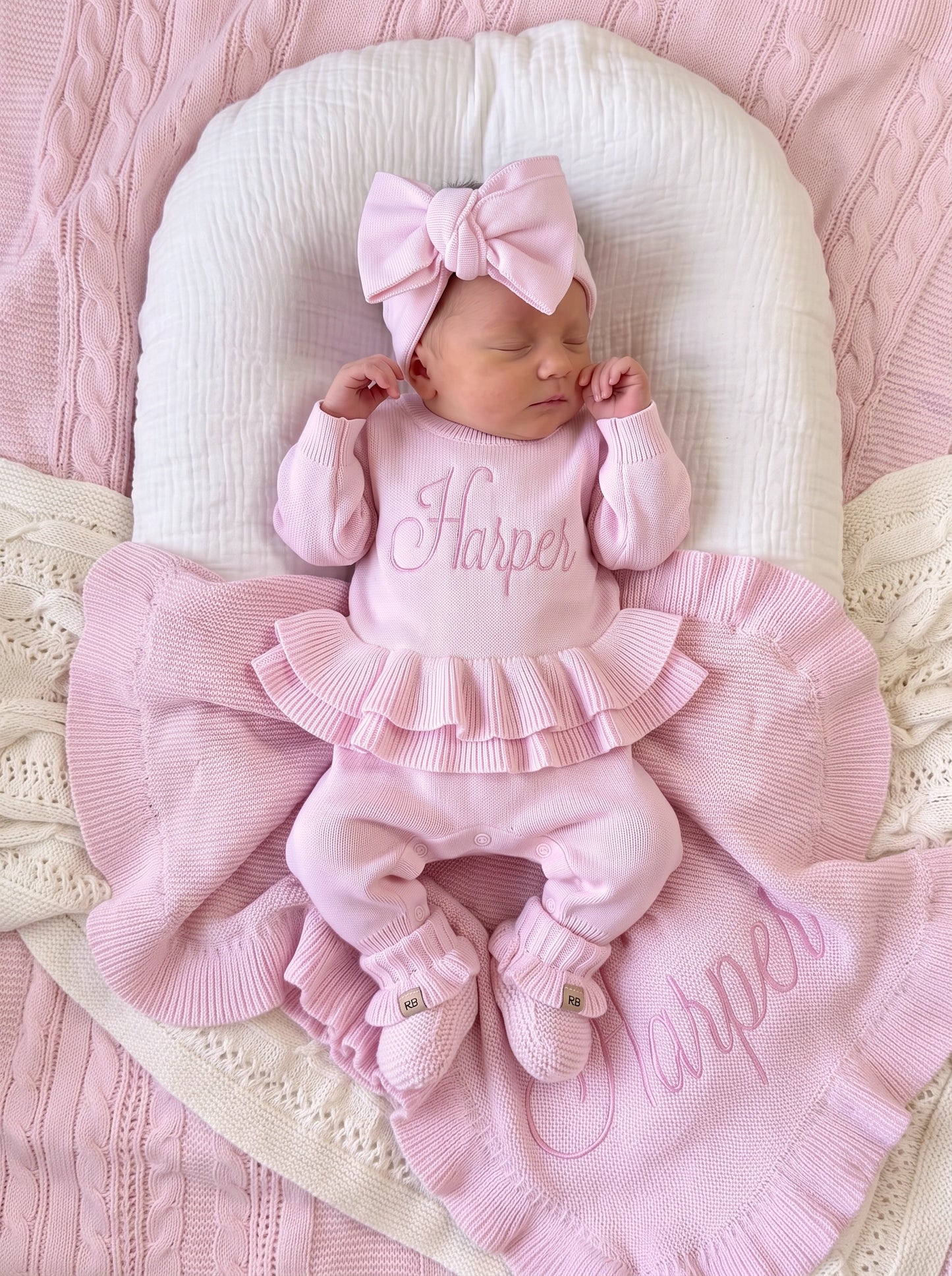 Newborn wearing a personalized petal pink Ballerina long romper with bow headwrap, resting on a matching ruffle knit blanket.