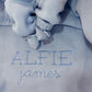 Close-up of Baby Blue classic knit booties resting on a personalized newborn blanket embroidered with the name Alfie James.
