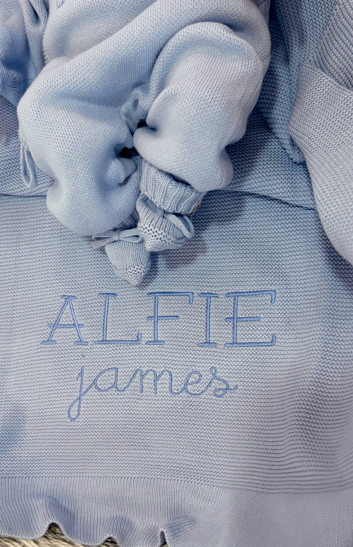 Close-up of Baby Blue classic knit booties resting on a personalized newborn blanket embroidered with the name Alfie James.