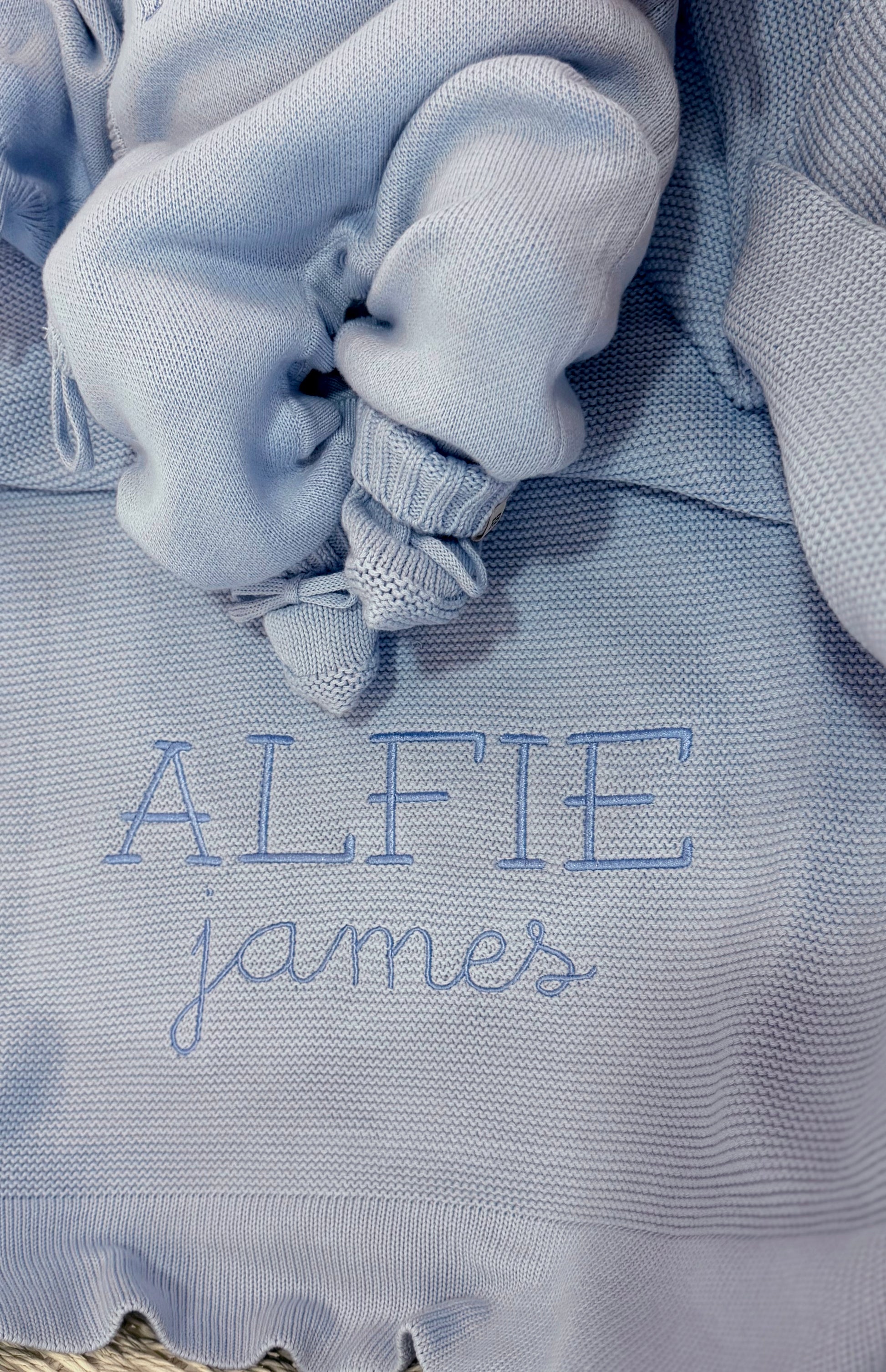 Close-up of Baby Blue classic knit booties resting on a personalized newborn blanket embroidered with the name Alfie James.
