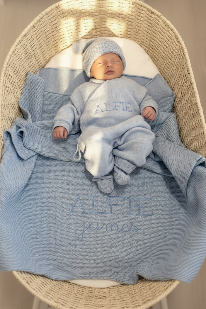 Sleeping newborn wearing the personalized Baby Blue Classic Knit long romper set in a woven bassinet with a matching Alfie blanket.