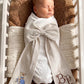 Swaddled newborn baby boy styled with a personalized striped bow sash featuring embroidered teddy bear and monogram detail in a bassinet for hospital coming home photos and newborn keepsake moments.