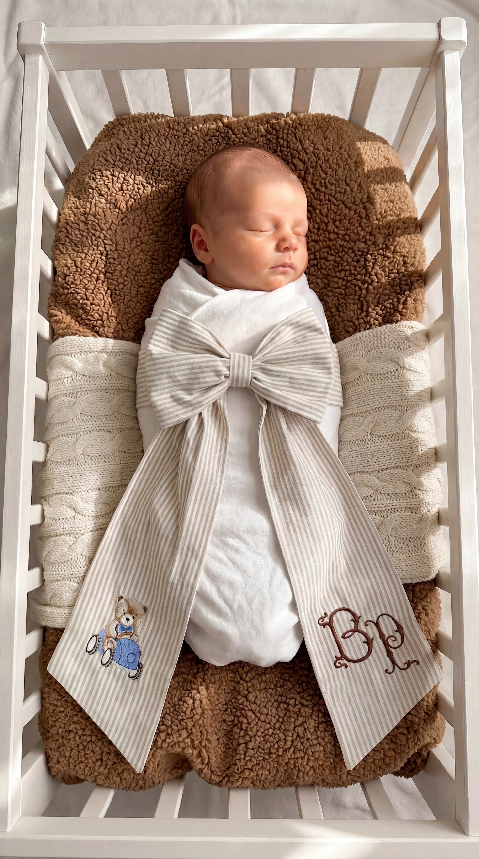 Swaddled newborn baby boy styled with a personalized striped bow sash featuring embroidered teddy bear and monogram detail in a bassinet for hospital coming home photos and newborn keepsake moments.