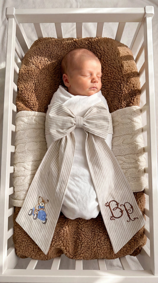 Swaddled newborn baby boy styled with a personalized striped bow sash featuring embroidered teddy bear and monogram detail in a bassinet for hospital coming home photos and newborn keepsake moments.