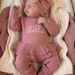 Sleeping newborn wearing the personalized Dusty Mauve Ballerina long romper with oversized bow and ruffle booties in a woven bassinet.