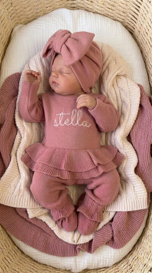 Sleeping newborn wearing the personalized Dusty Mauve Ballerina long romper with oversized bow and ruffle booties in a woven bassinet.