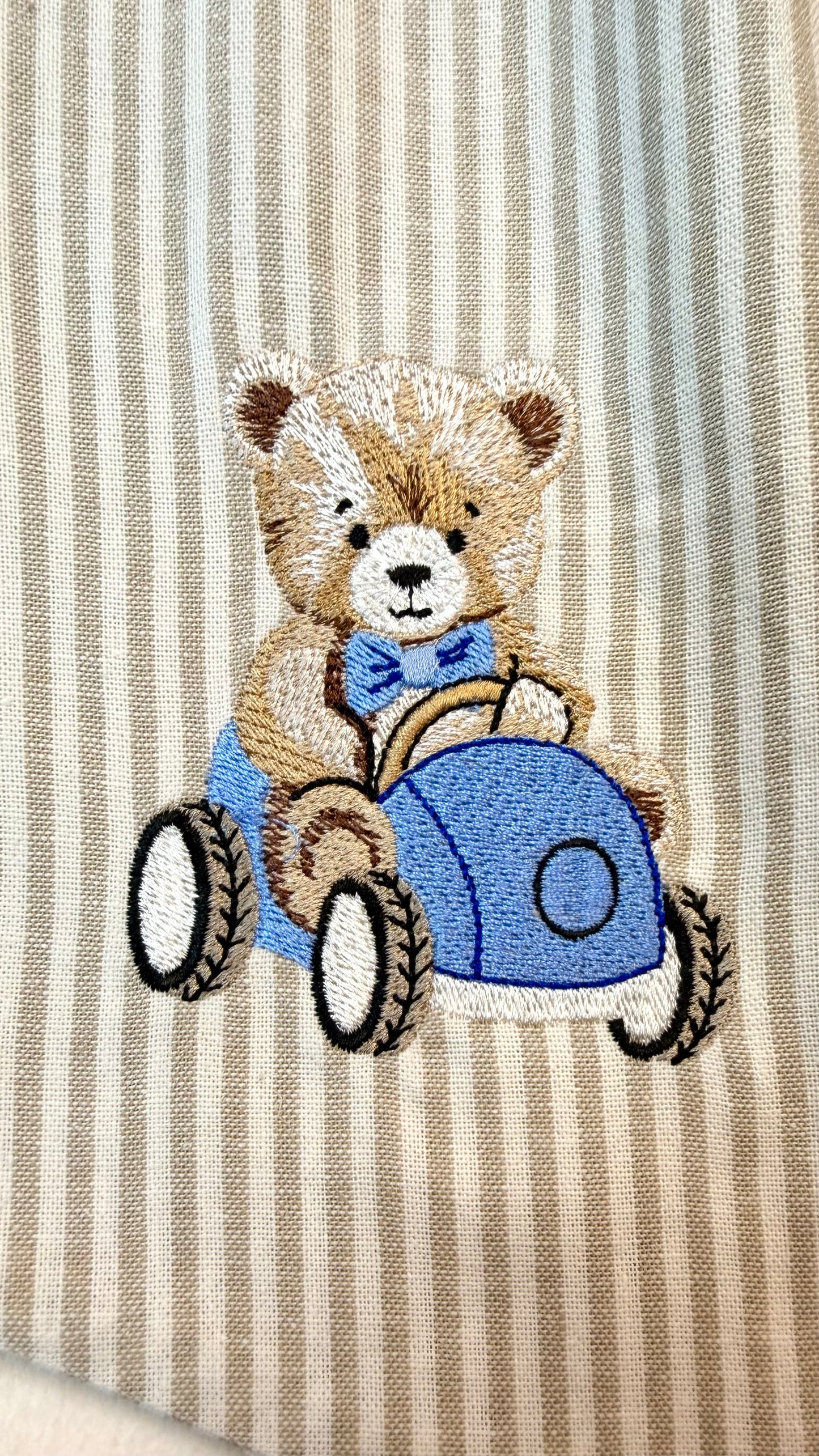 Vintage-style teddy bear embroidery driving a blue car on soft striped knit fabric for a personalized baby boy outfit or newborn keepsake clothing detail.