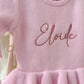 Detailed view of embroidered name on personalized Petal Pink ballerina baby romper showing soft knit fabric and ruffle trim