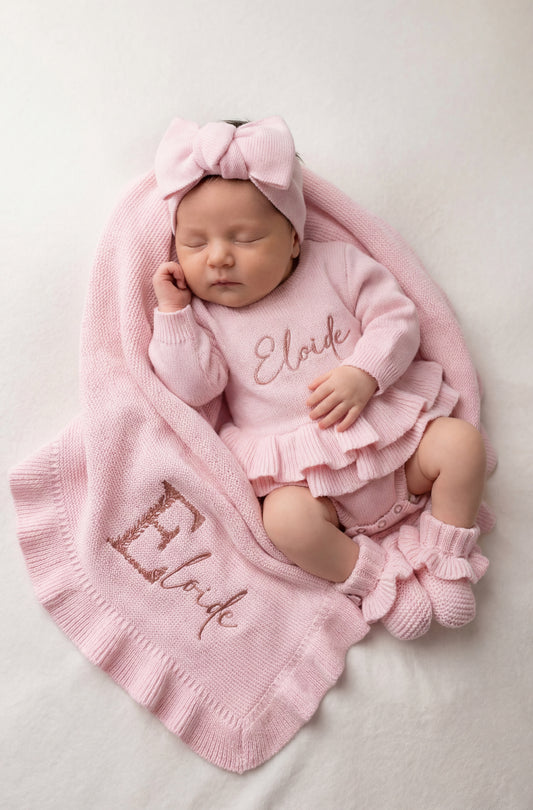 Newborn baby wearing personalized Petal Pink outfit set with embroidered name, including romper, bow, booties, and blanket