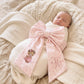 Swaddled newborn baby girl wearing a personalized pink bow sash with embroidered teddy bear and monogram detail, styled for hospital coming home photos on soft neutral knit blankets.