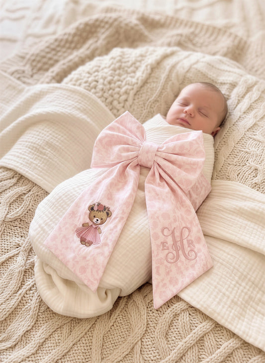 Swaddled newborn baby girl wearing a personalized pink bow sash with embroidered teddy bear and monogram detail, styled for hospital coming home photos on soft neutral knit blankets.