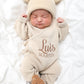 Newborn baby wearing classic knit wrap romper in soft khaki with personalized embroidery, styled with knit hat and teddy bear