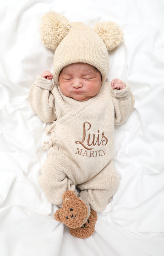 Newborn baby wearing classic knit wrap romper in soft khaki with personalized embroidery, styled with knit hat and teddy bear