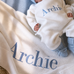 Newborn in the personalized Warm White Kimono long romper with Baby Blue hat and booties beside a matching blanket embroidered with Archie.