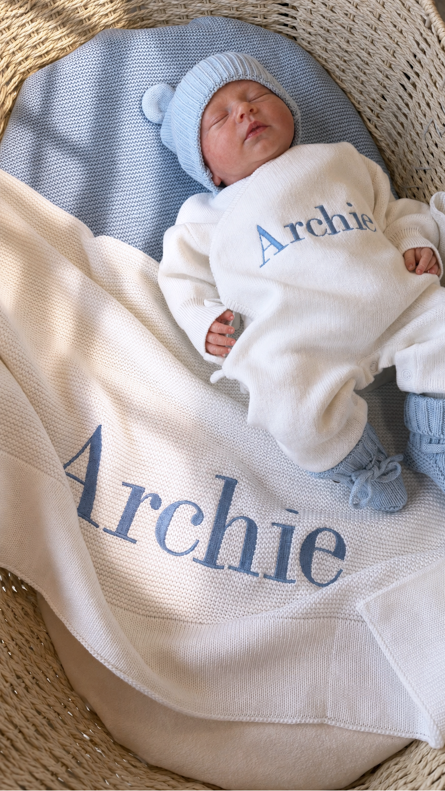 Newborn in the personalized Warm White Kimono long romper with Baby Blue hat and booties beside a matching blanket embroidered with Archie.