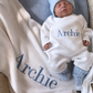 Sleeping newborn wearing the personalized Warm White and Baby Blue Kimono long romper with bear-ear hat and booties beside an Archie embroidered blanket.