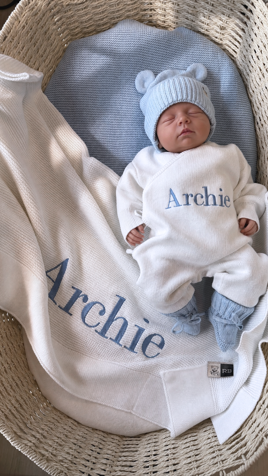 Sleeping newborn wearing the personalized Warm White and Baby Blue Kimono long romper with bear-ear hat and booties beside an Archie embroidered blanket.