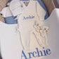 Complete personalized Warm White Kimono long romper set with matching hat, booties and Archie embroidered blanket displayed in a woven bassinet.