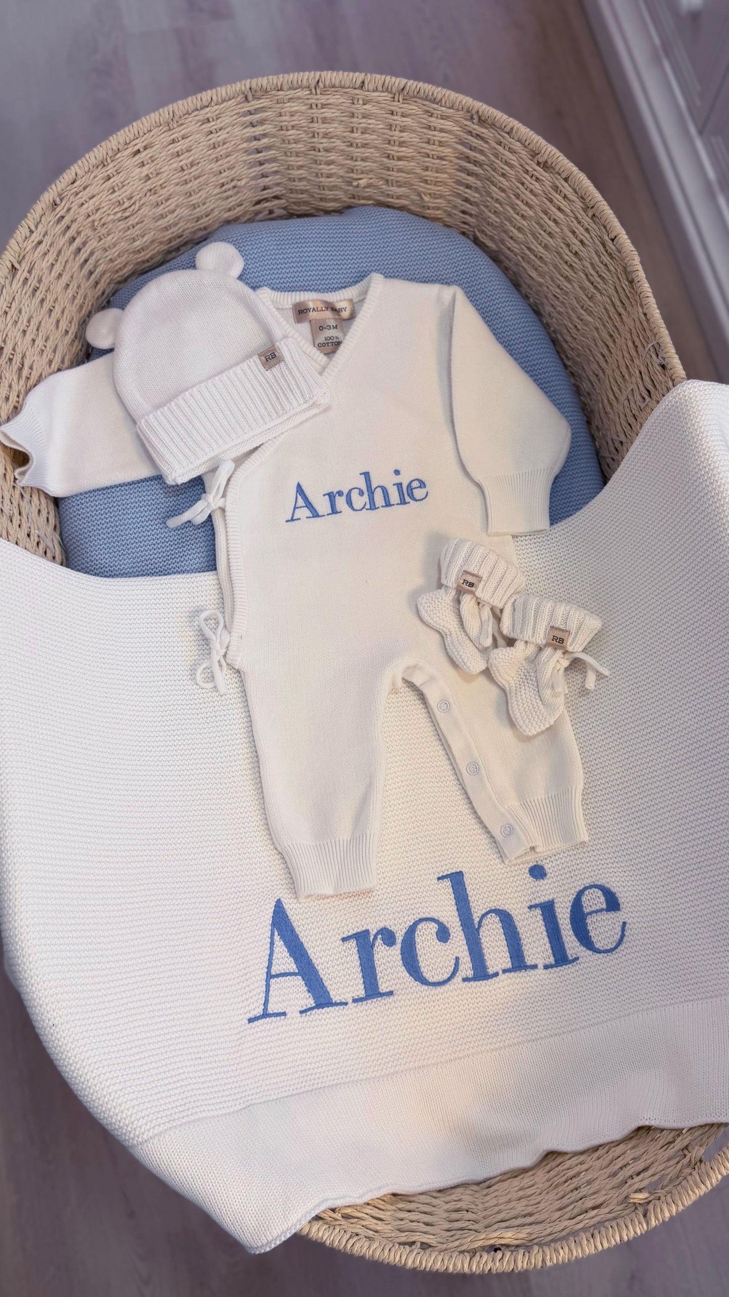 Complete personalized Warm White Kimono long romper set with matching hat, booties and Archie embroidered blanket displayed in a woven bassinet.