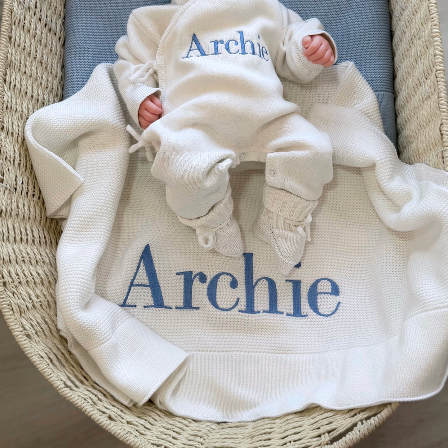 Overhead flat lay of the Warm White Kimono long romper, bear-ear hat and booties on a personalized Archie knit blanket.